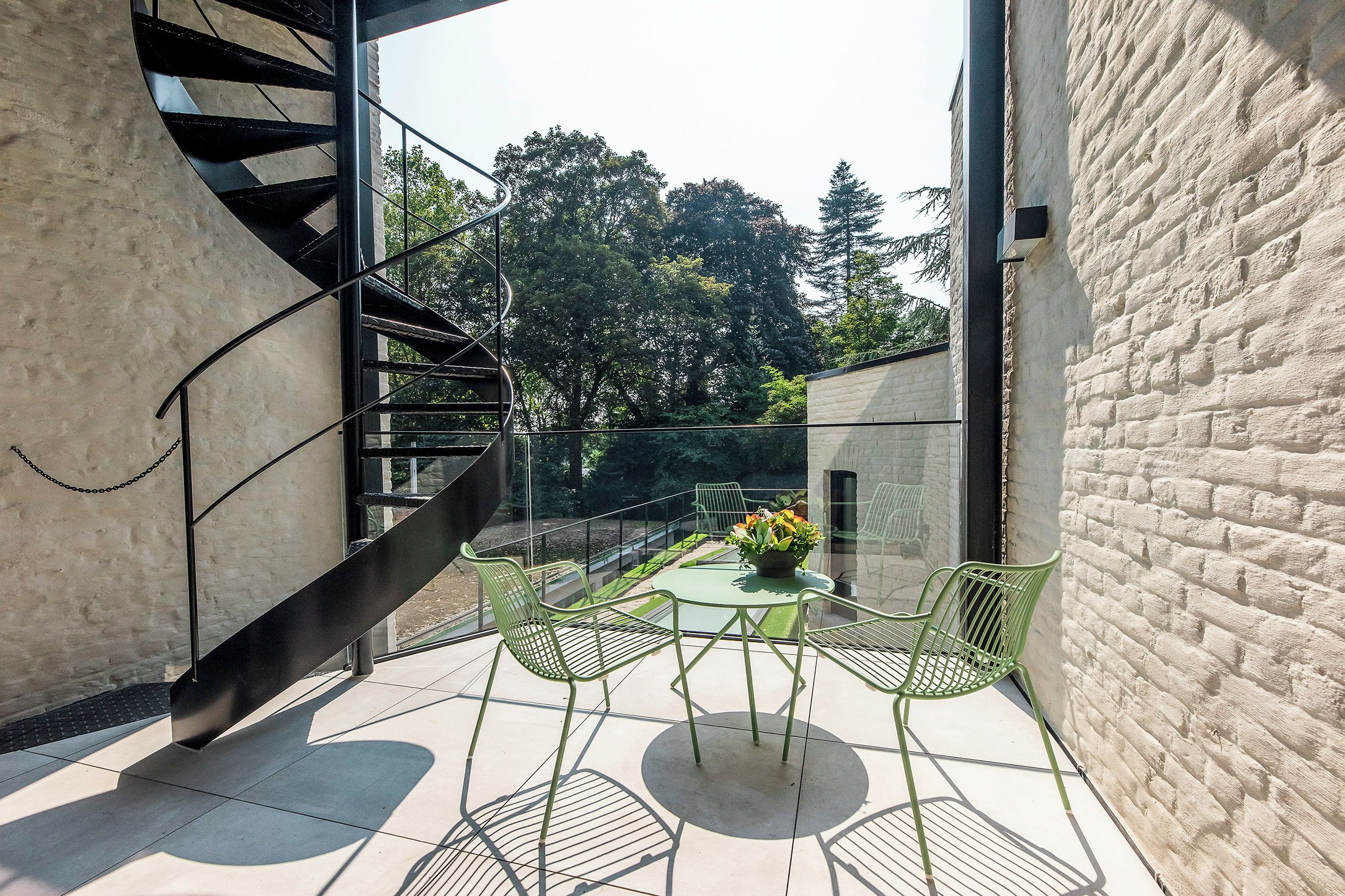 A modern outdoor seating area with a spiral staircase, a glass balustrade, and a view of trees.
