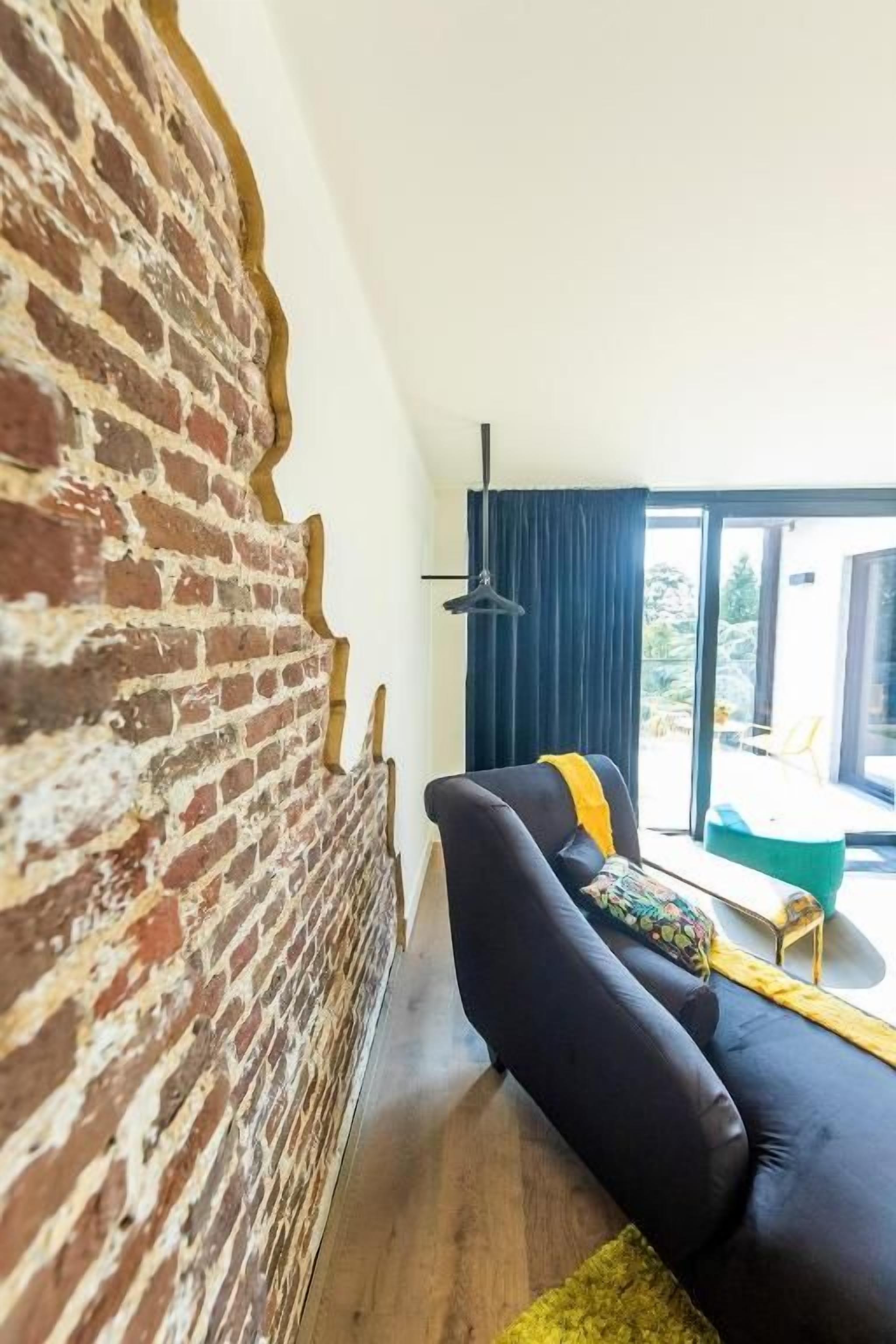 A room with a brick wall, a black sofa, a yellow throw, and a view through a window.