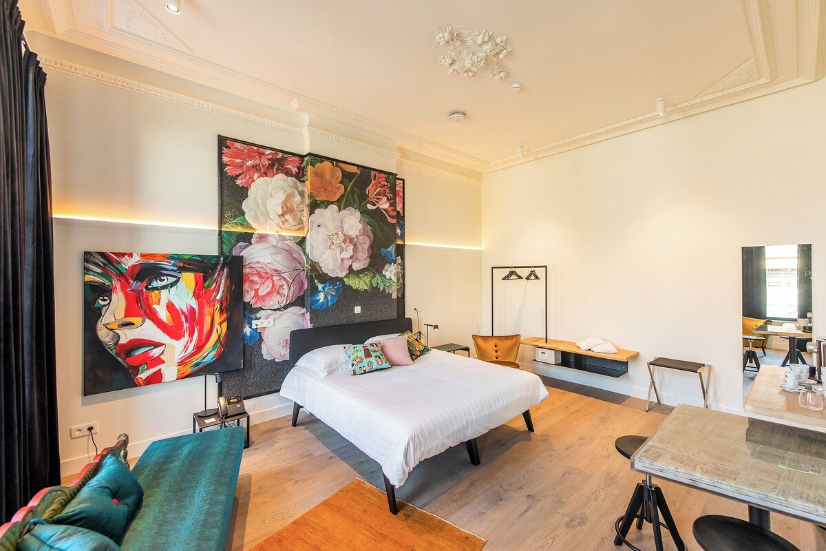 A modern hotel room with a large floral mural, a bed with white linens, a teal armchair, and a dining area with a glass table.