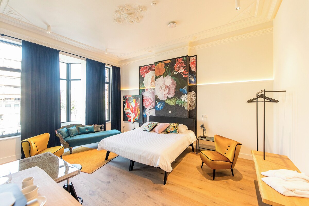 A hotel room with a bed, a sofa, a chair, a desk, and a large floral artwork on the wall.