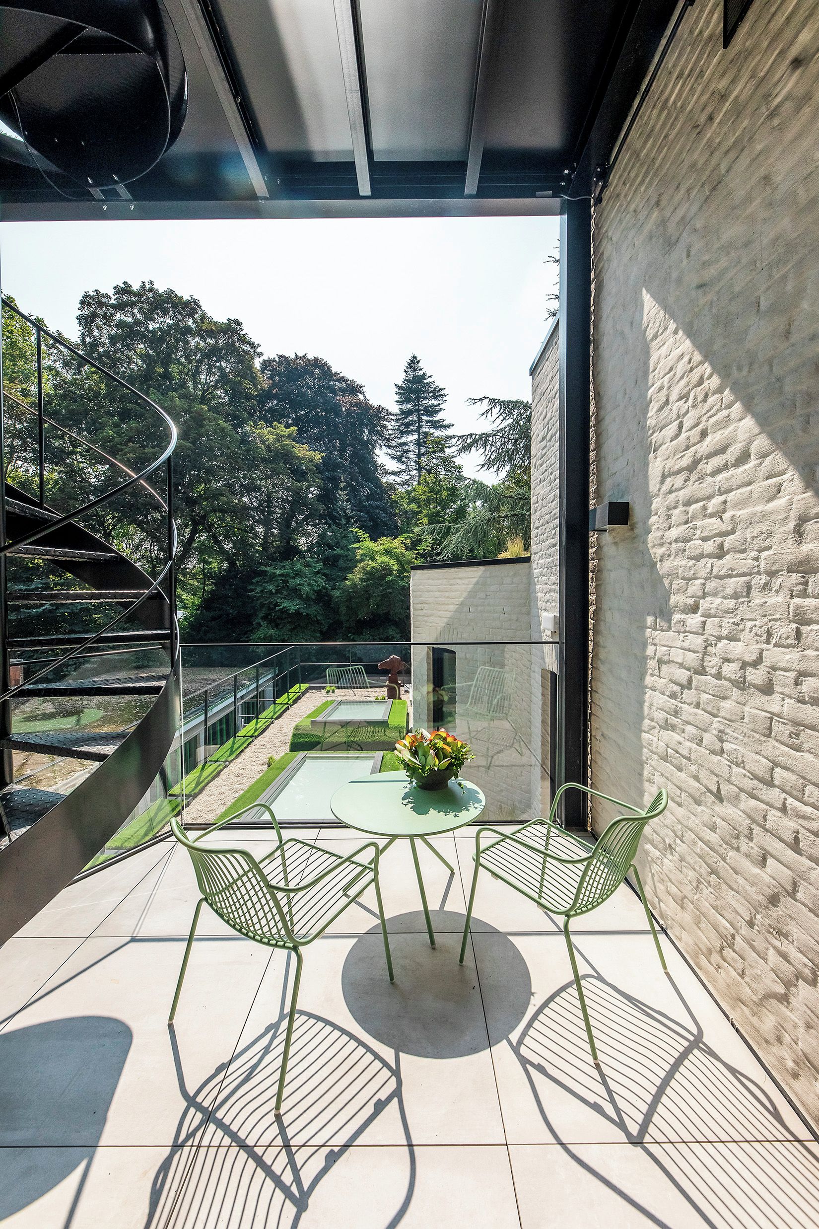 An outdoor patio with a spiral staircase, a round table with chairs, and a view of trees and a building in the background.