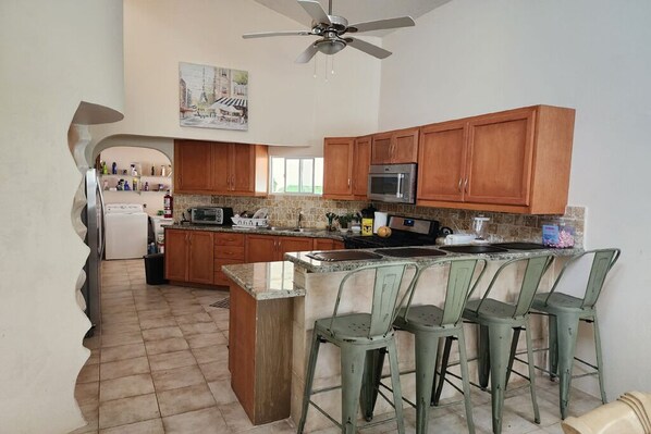 A kitchen with wooden cabinets, a microwave, a refrigerator, and a bar area with stools.