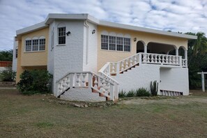 A two-story house with a white exterior, a balcony with white balustrades, and a small staircase leading to the entrance. - Anguilla Vacation Home – Cradle Grove (Little Harbour)