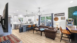 Een moderne woonkamer met een bank, salontafel en een barhoek. In de achtergrond een keuken met witte kasten en een eethoek met oranje stoelen.