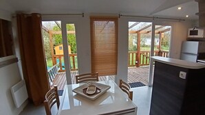 A compact kitchen with a dining area, a small refrigerator, and a view of a wooden deck and garden. - Mobil-home 6 pers - Sea & nature -camping 3* familial et calme - Plot XXL (Arces)