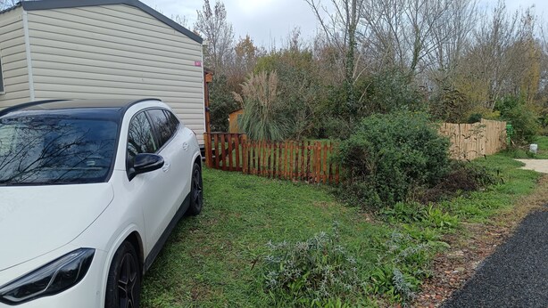 A white car parked in a grassy area with a wooden fence and a house in the background. - Mobil-home 6 pers - Sea & nature -camping 3* familial et calme - Plot XXL (Arces)
