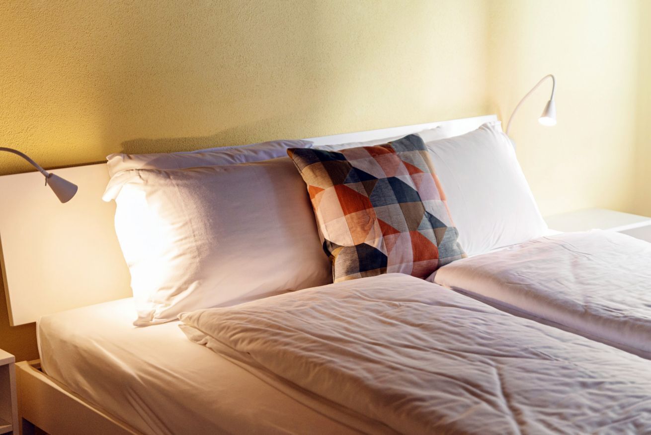 A neatly made bed with white bedding and a colorful geometric pillow, a bedside lamp, and a plain wall.