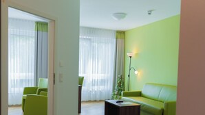 A modern living room with a green sofa, a coffee table, and a lamp. There is a door leading to another room, and a plant is visible in the corner. - ALEMBRA - Wein Style Hotel (Trier)