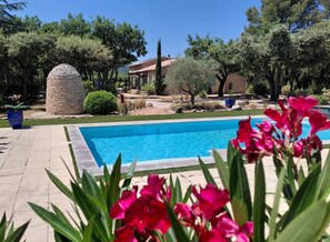 A swimming pool with a stone structure nearby, surrounded by trees and a building in the background. - mas val-chenaie (Gordes)