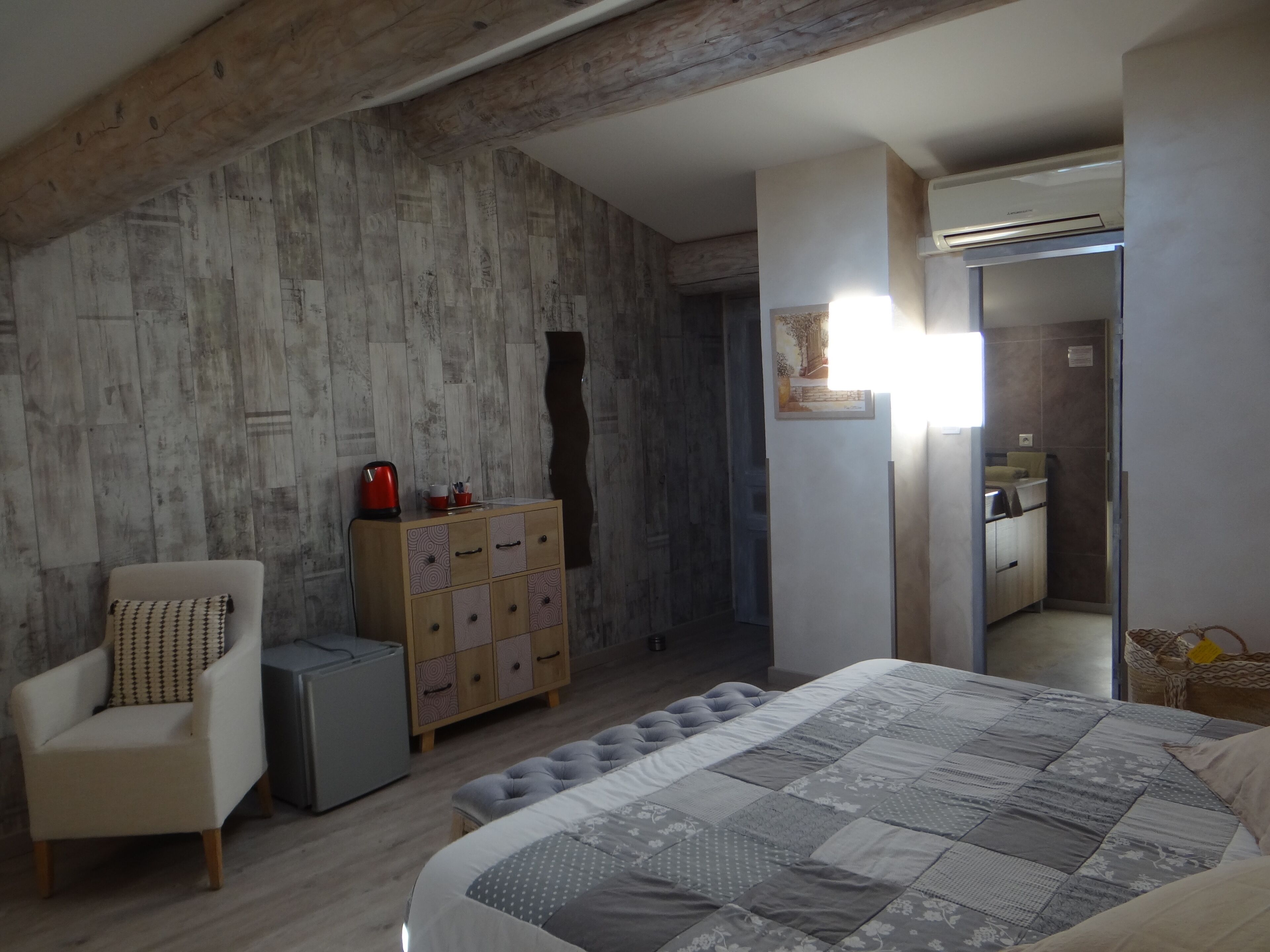 Ein modernes Schlafzimmer mit Holzboden, einem Bett mit grauer Tagesdecke, einem Schrank mit Schubladen, einem Stuhl und einem kleinen Tisch mit Wasserkocher.