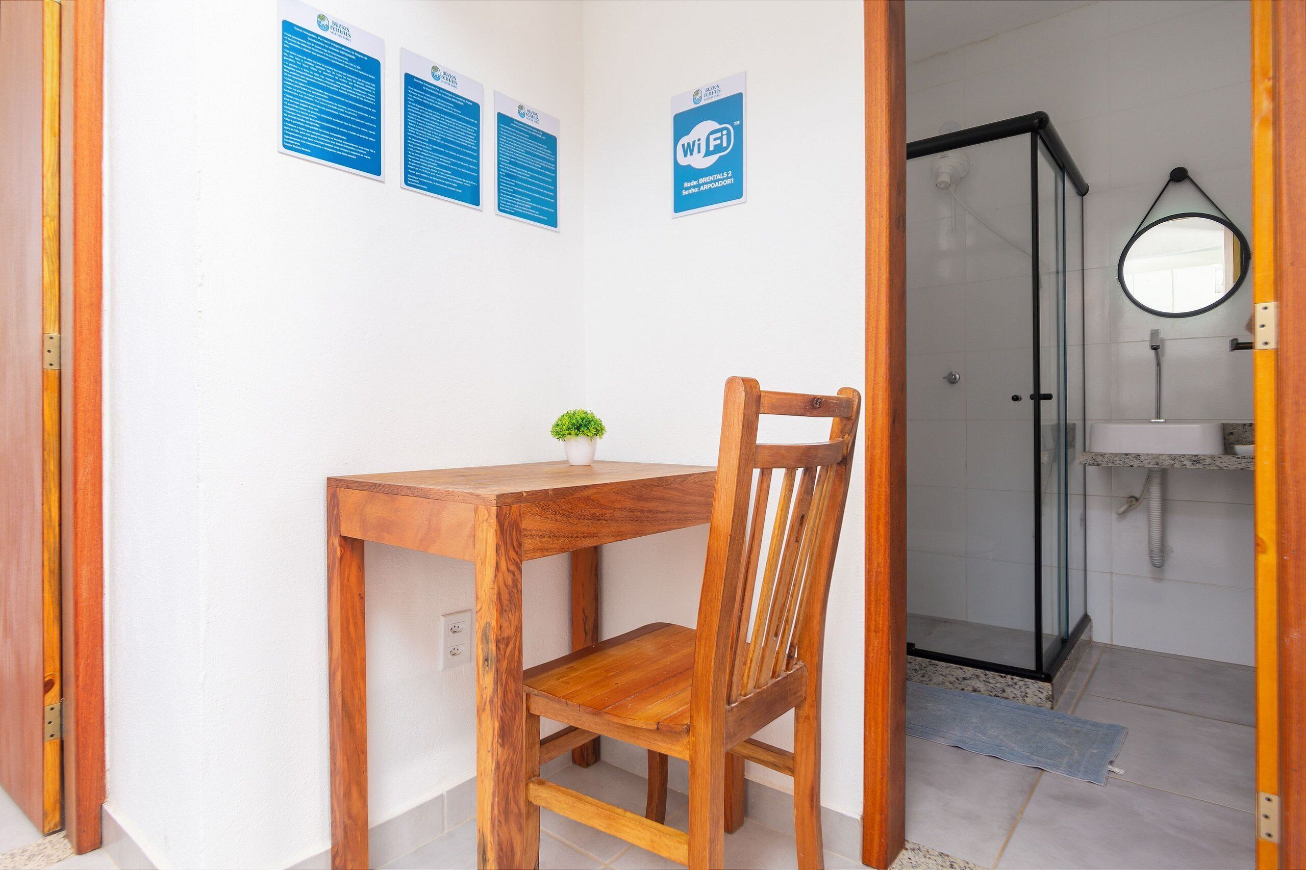 Quarto com mesa e cadeira de madeira, uma pequena planta em vaso, box de vidro para o chuveiro e espelho na parede.
