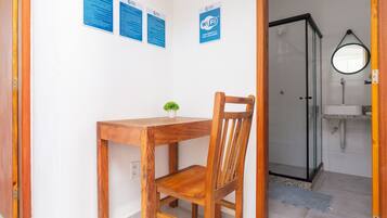 Quarto com mesa e cadeira de madeira, uma pequena planta em vaso, box de vidro para o chuveiro e espelho na parede.