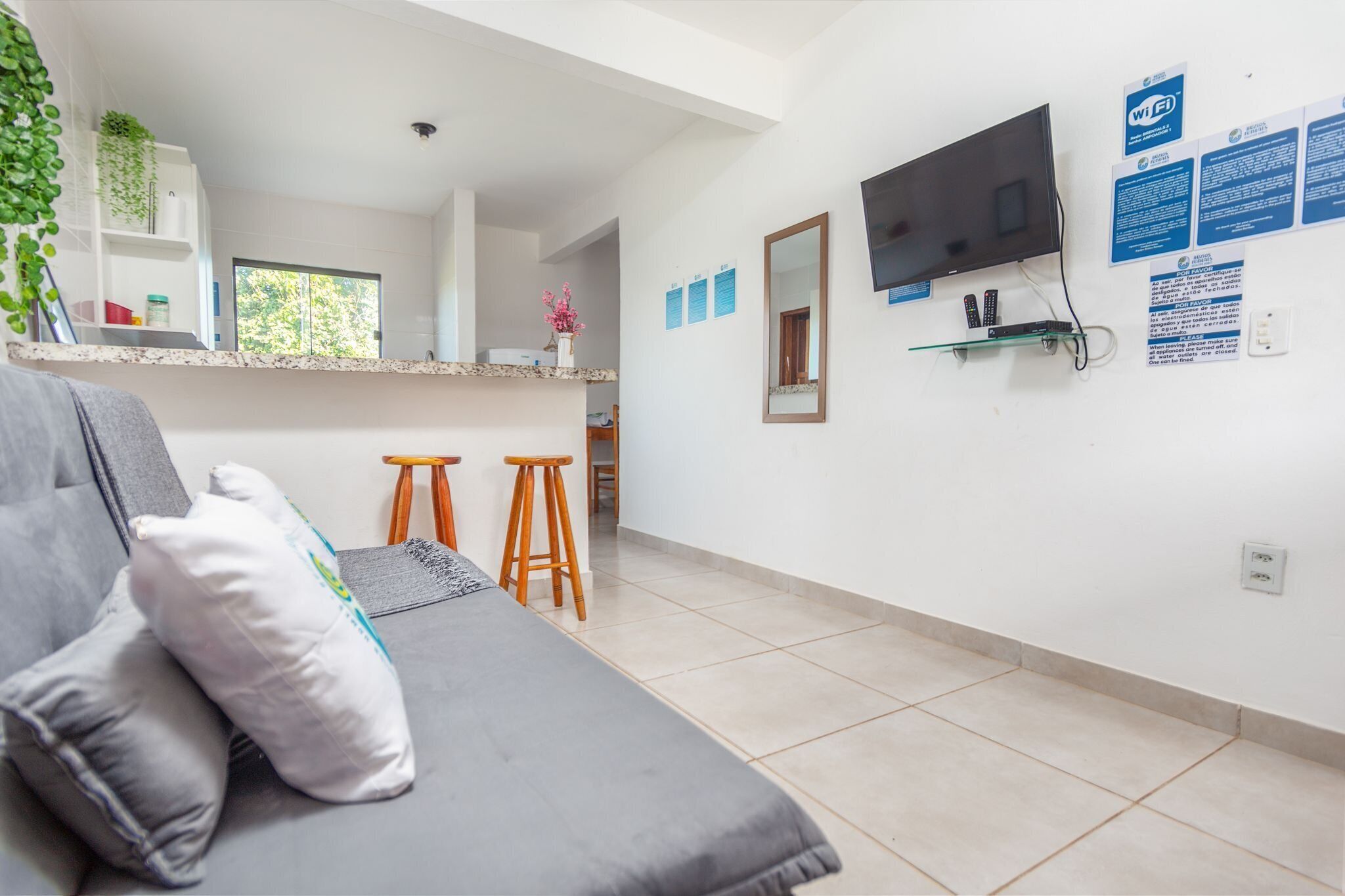 Une pièce moderne avec un canapé, deux tabourets en bois, une télévision et un coin cuisine à l’arrière-plan.