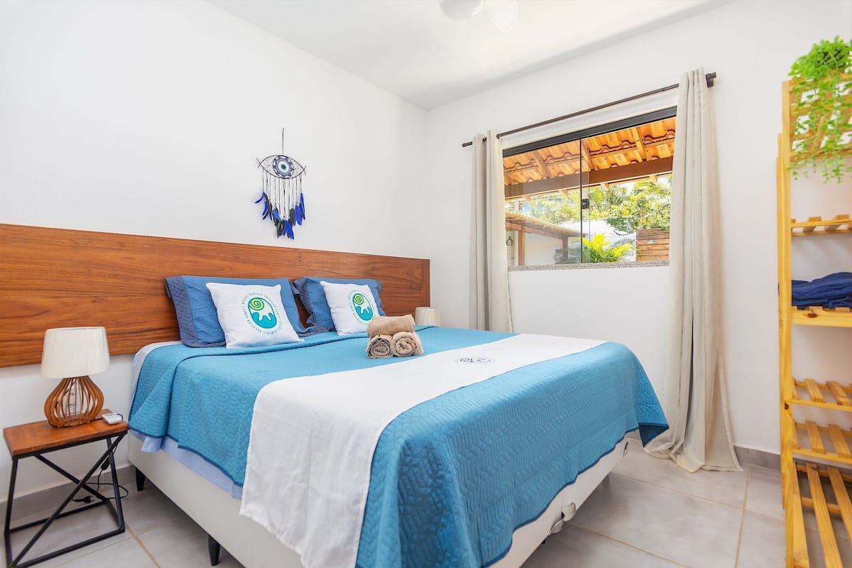 A neatly made bed with a blue and white bedspread, a wooden headboard, a bedside table with a lamp, and a window with a view of greenery.