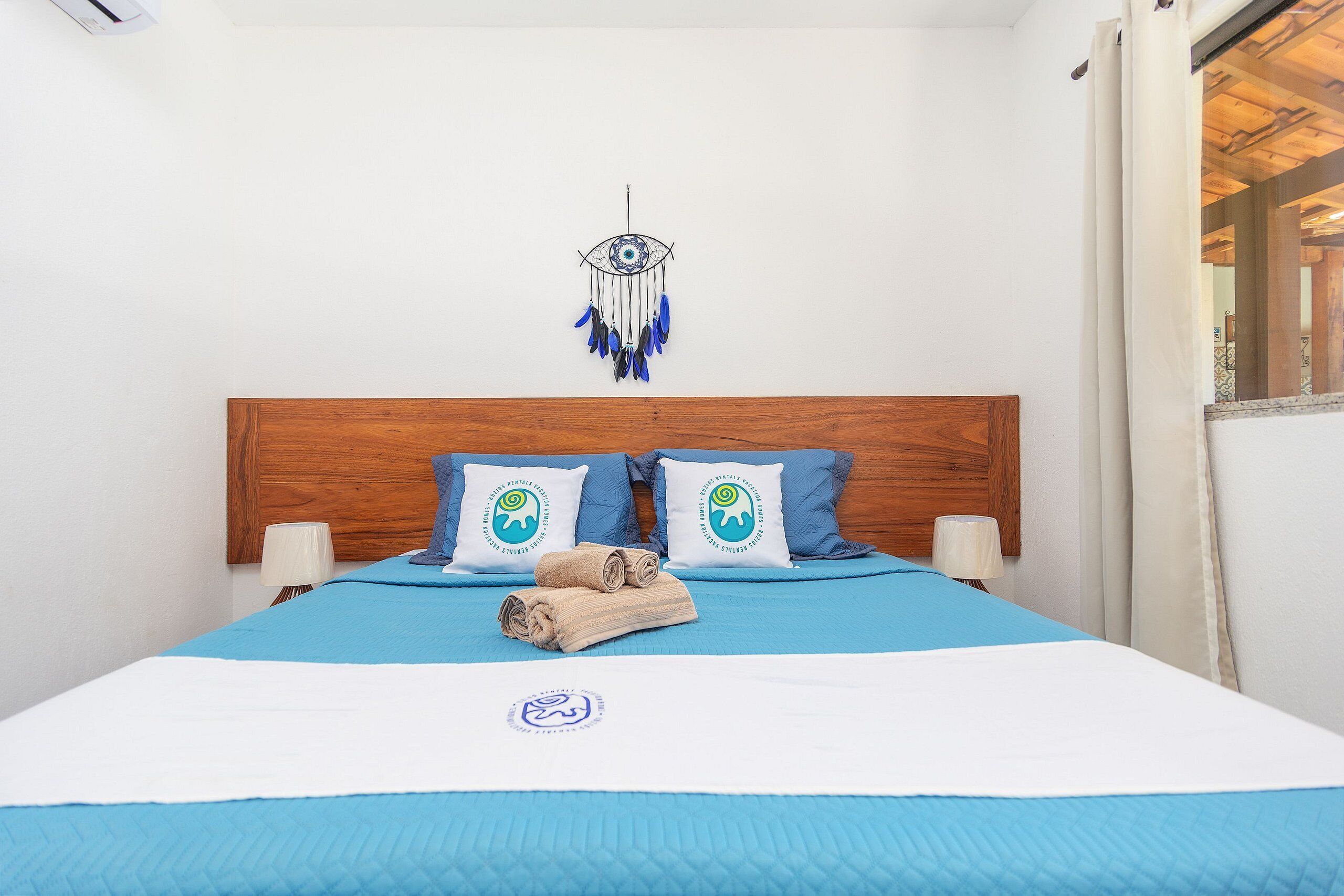 A neatly made bed with blue and white bedding, two pillows with a logo, a wooden headboard, and a wall-mounted dreamcatcher.