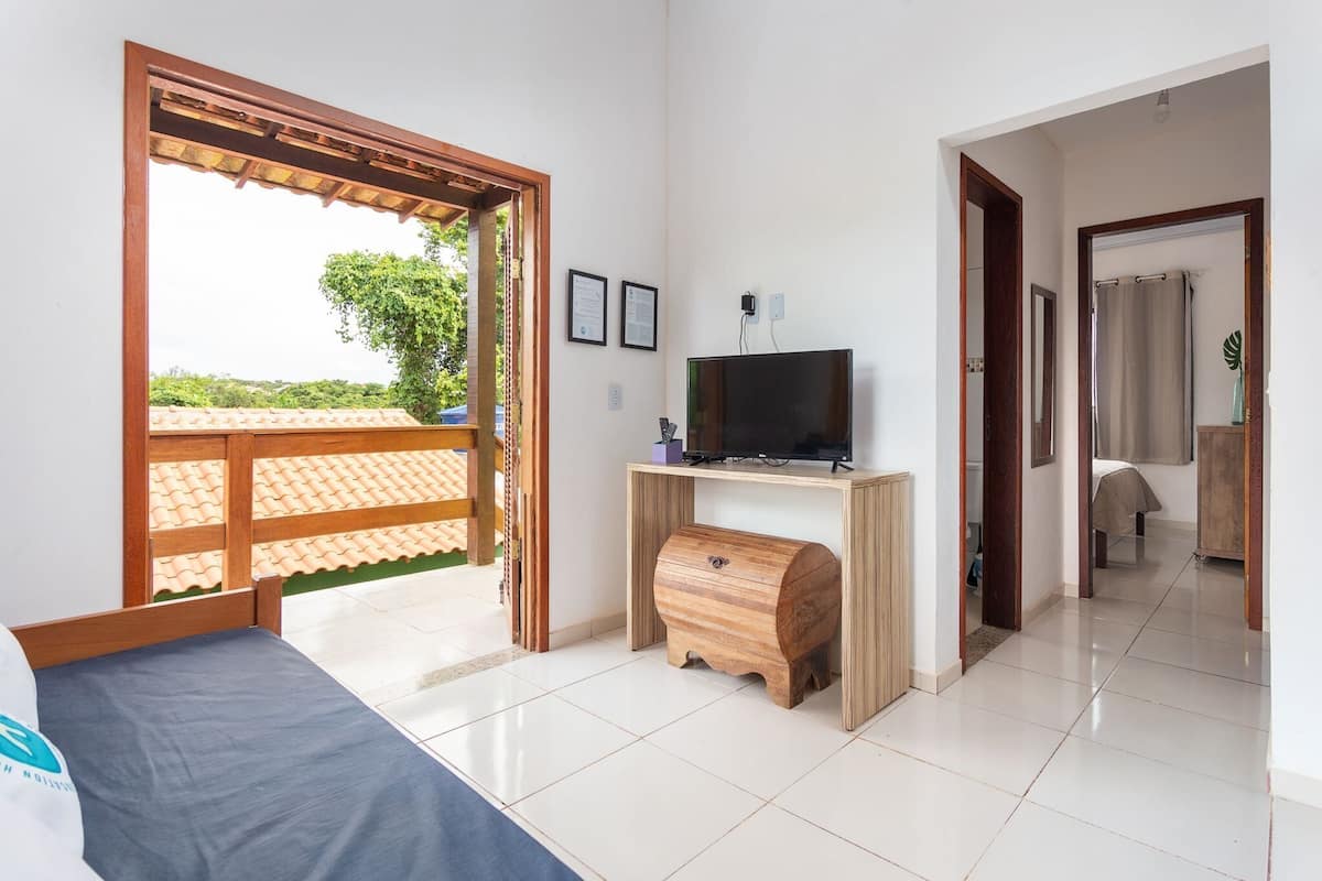 A bedroom with a bed, a TV on a wooden stand, a wooden chest, and a view of a tiled patio.