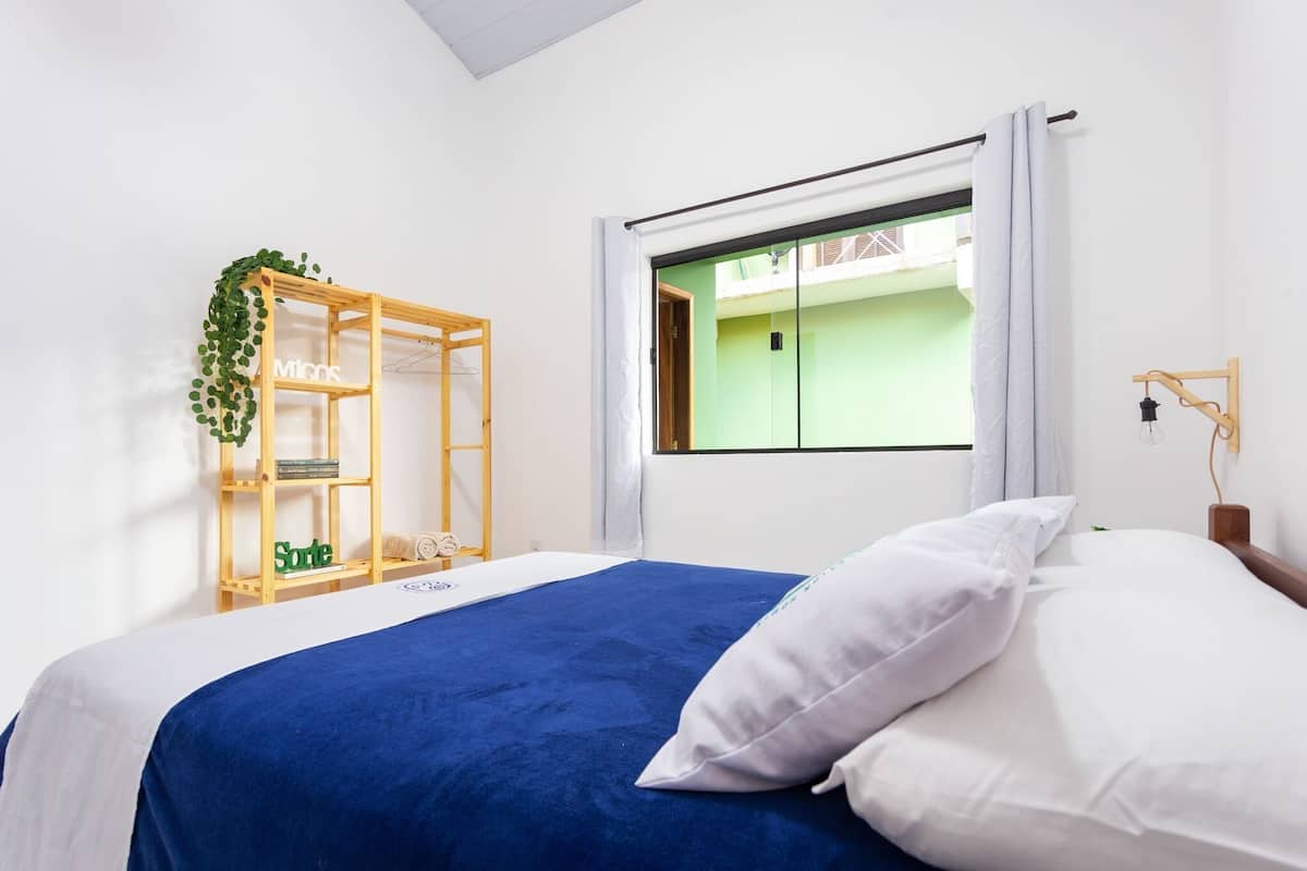 A bedroom with a bed, a window with curtains, a wooden shelf with greenery, and a wall-mounted lamp.