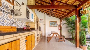 Un espacio al aire libre techado con mesa y bancos de madera, mostrador de ladrillo con armarios y suelo de baldosas.