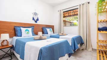 A hotel room with two beds, a wooden headboard, a nightstand with a lamp, a window with curtains, and a shelf with plants.