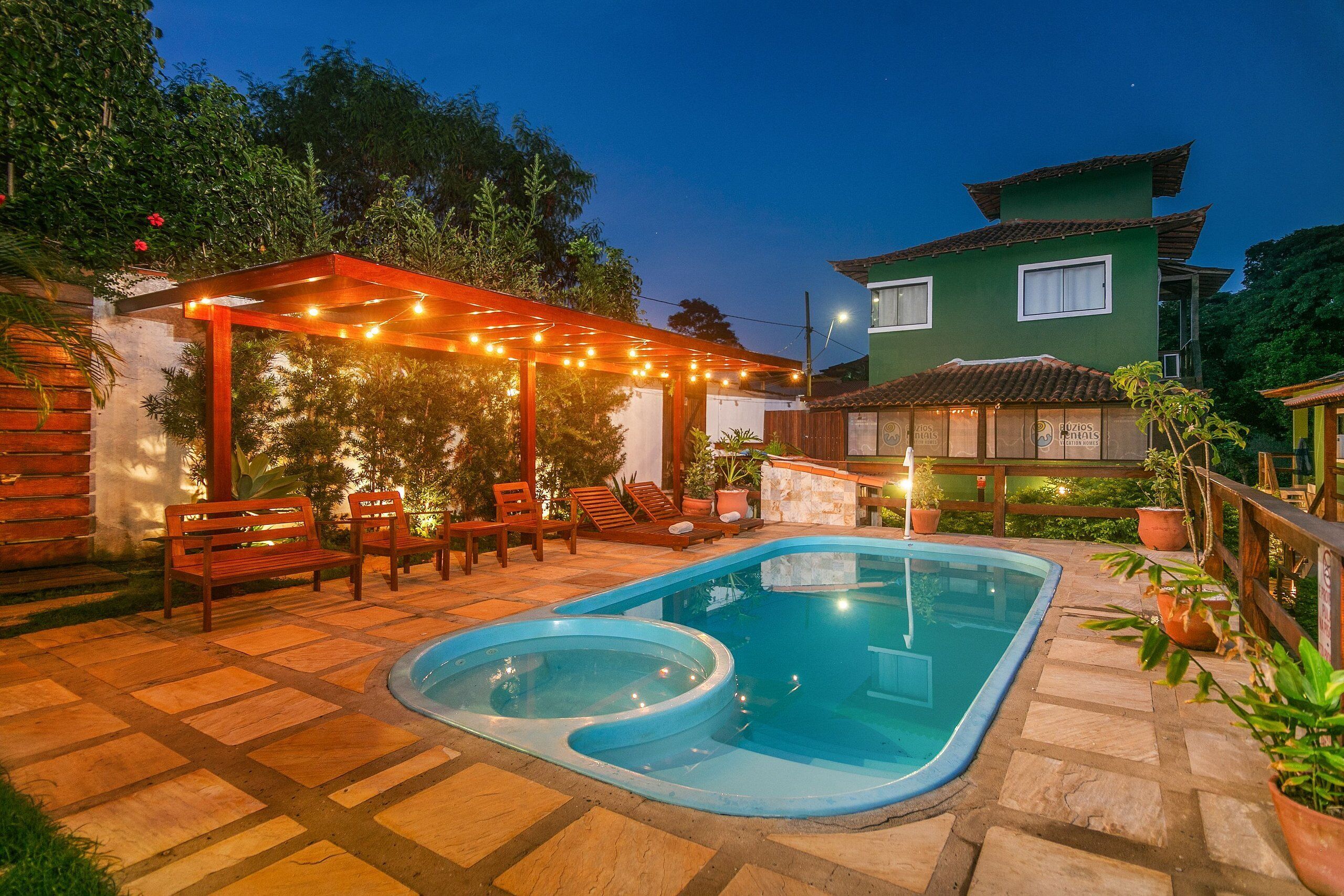 Piscina com deck de madeira e espreguiçadeiras, cercada por plantas em vasos e uma pérgola com luzes de cordão.