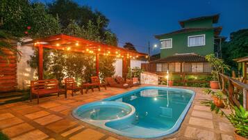 Piscina com deck de madeira e espreguiçadeiras, cercada por plantas em vasos e uma pérgola com luzes de cordão.