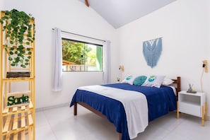 A bedroom with a bed, a nightstand, a window with curtains, a wall-mounted macrame, and a wooden shelf with plants.