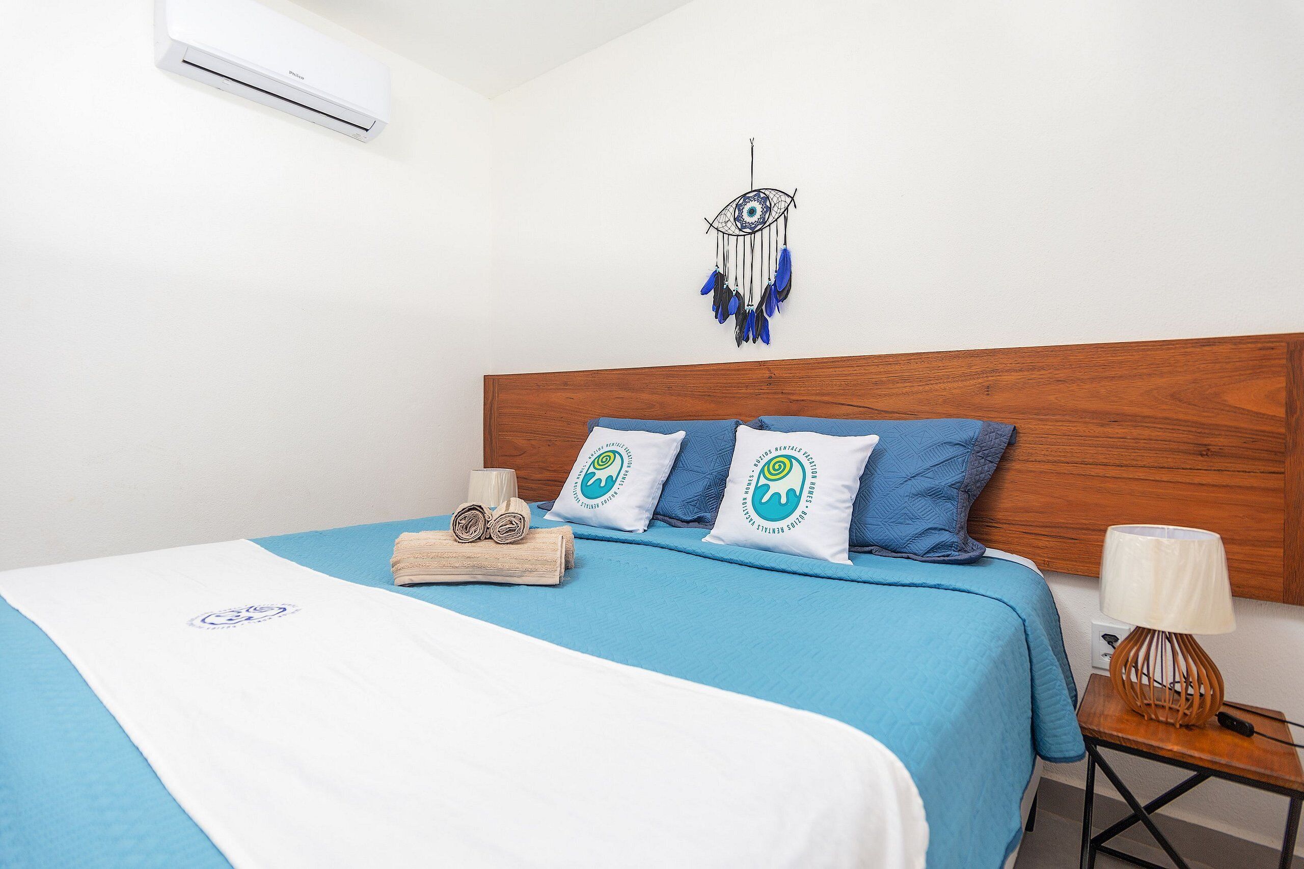 A neatly made bed with a blue bedspread, two pillows with a circular logo, a wooden headboard, a dreamcatcher, a small wooden nightstand with a lamp, and an air conditioning unit on the wall.