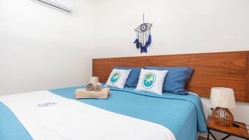 A neatly made bed with a blue bedspread, two pillows with a circular logo, a wooden headboard, a dreamcatcher, a small wooden nightstand with a lamp, and an air conditioning unit on the wall.