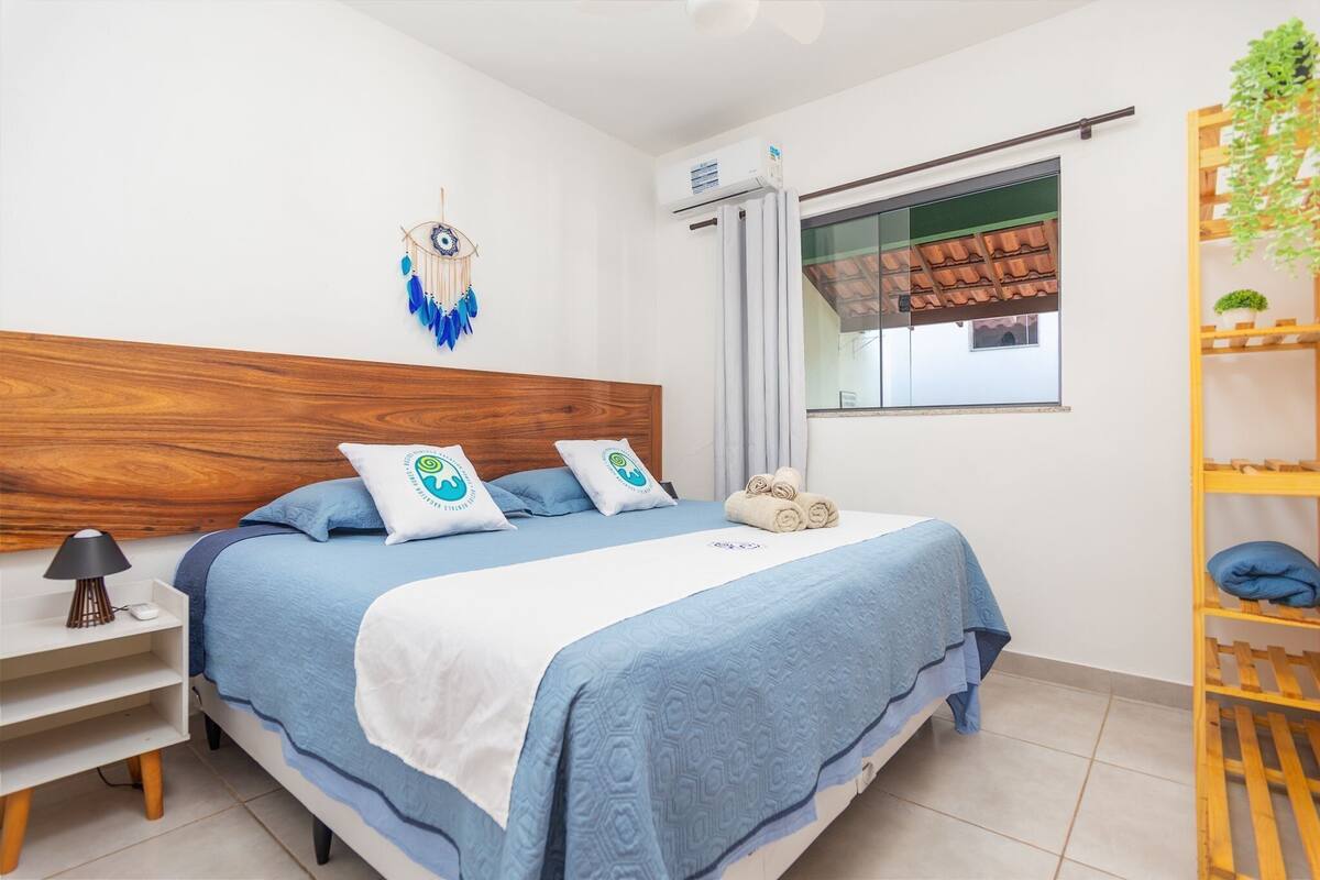 Une chambre avec une tête de lit en bois, un lit avec une literie bleue, une table de chevet avec une lampe, un attrape-rêves, un appareil de climatisation, une fenêtre avec vue et une étagère en bois avec des plantes.