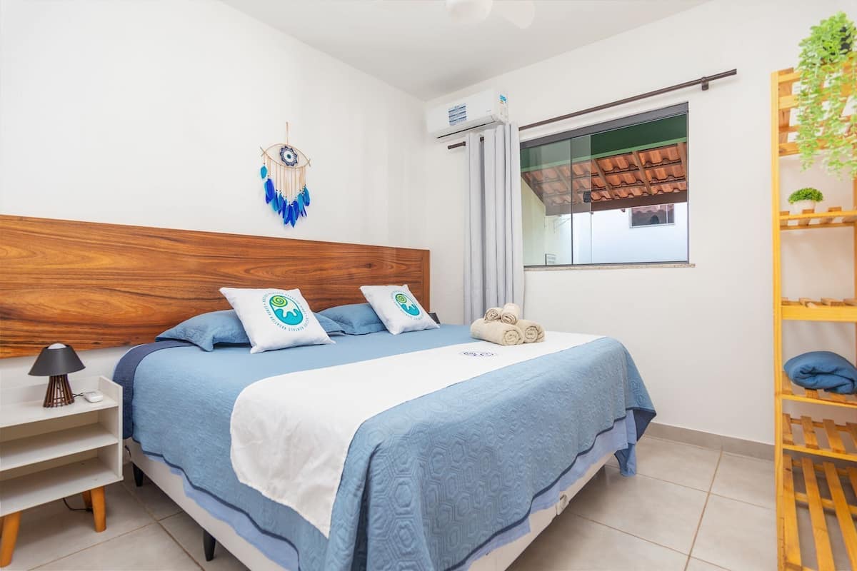 A bedroom with a wooden headboard, a bed with blue bedding, a bedside table with a lamp, a dreamcatcher, an air conditioning unit, a window with a view, and a wooden shelf with plants.