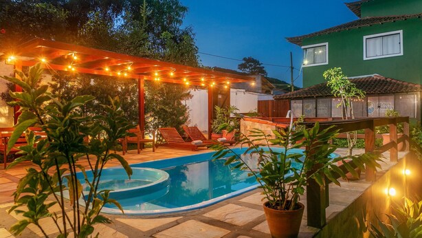 Una piscina con una pérgola y luces colgantes, rodeada de plantas en macetas y un invernadero.