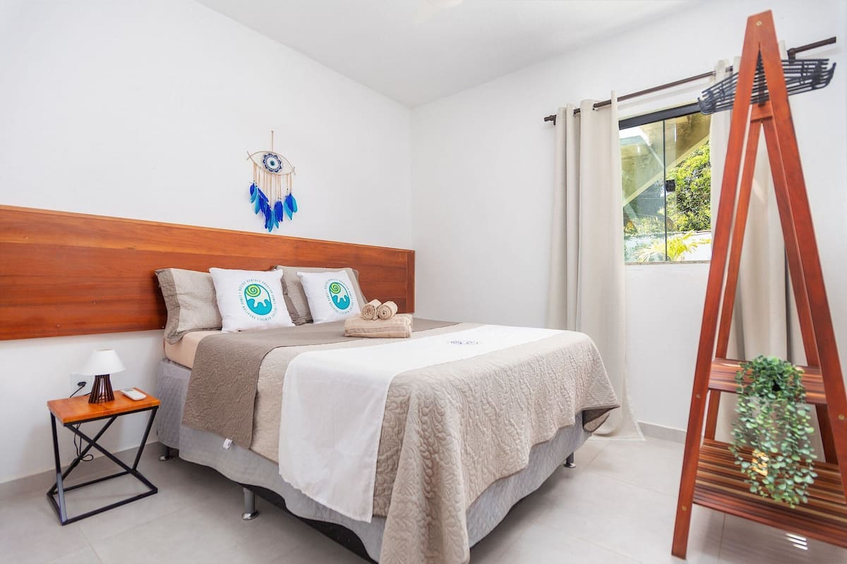 A bedroom with a wooden headboard, a bed with a beige blanket, a bedside table with a lamp, and a window with curtains.