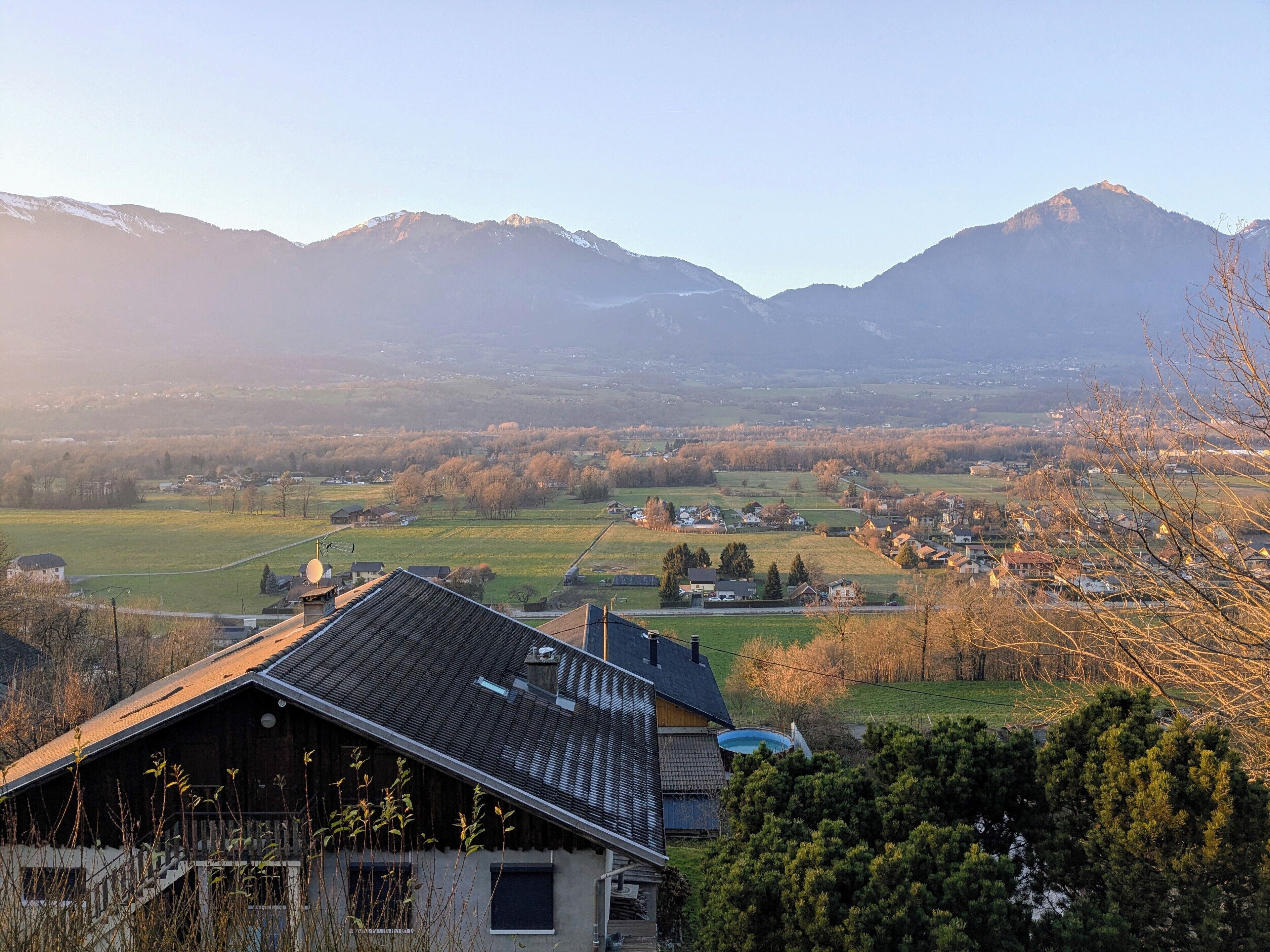 A rural landscape with mountains in the background, a house with a sloped roof, and a clear sky.