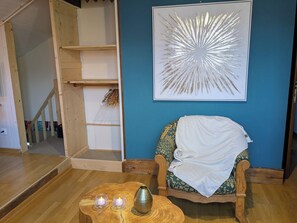 Ein Zimmer mit Holzboden, ein Stuhl mit Blumenkissen, ein kleiner Holztisch mit Vase und Kerzen sowie ein großes abstraktes Gemälde an der Wand.
