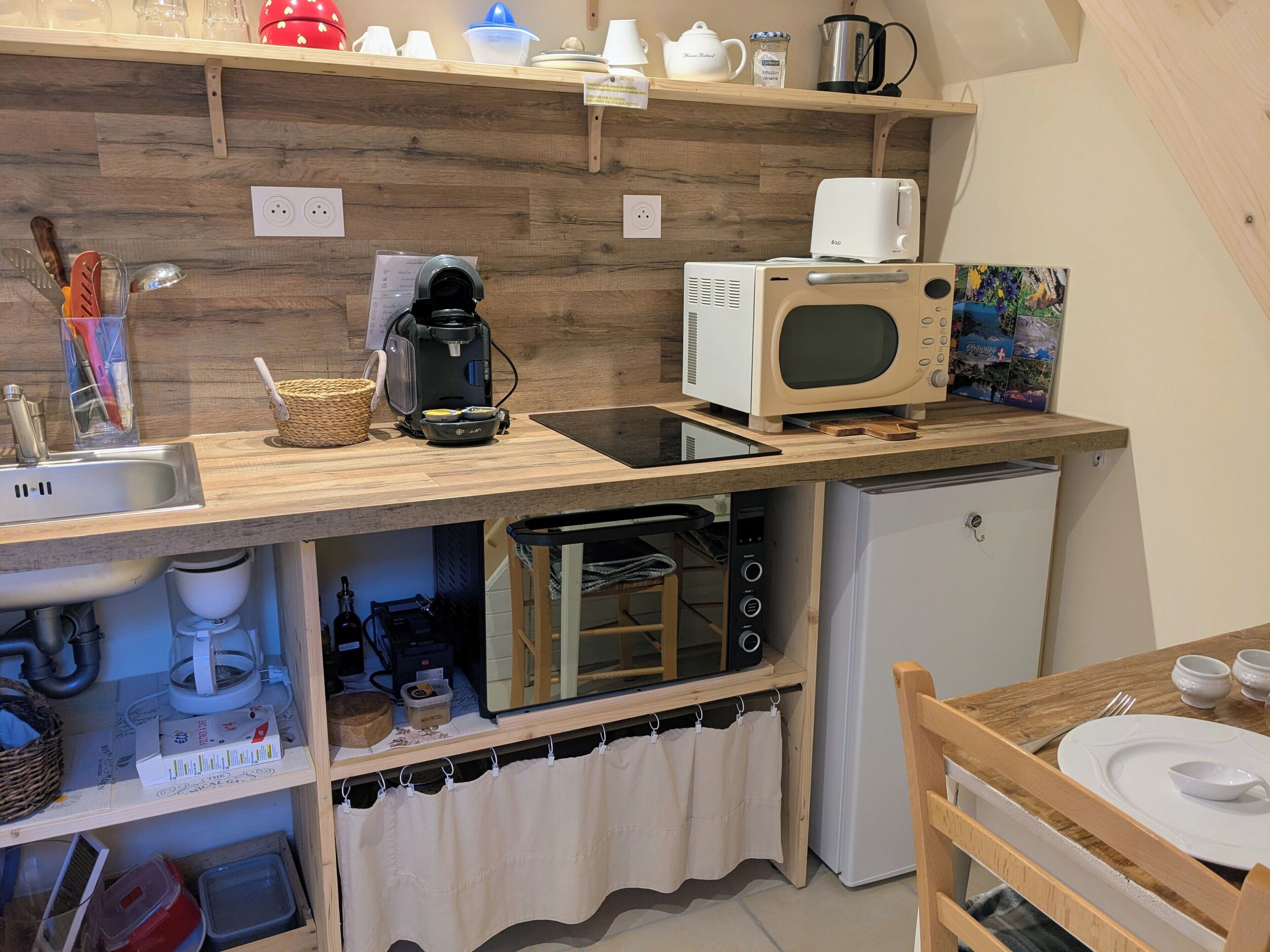 Una cocina compacta equipada con microondas, cafetera y un refrigerador pequeño. Hay repisas de madera sobre la encimera con diversos utensilios de cocina.