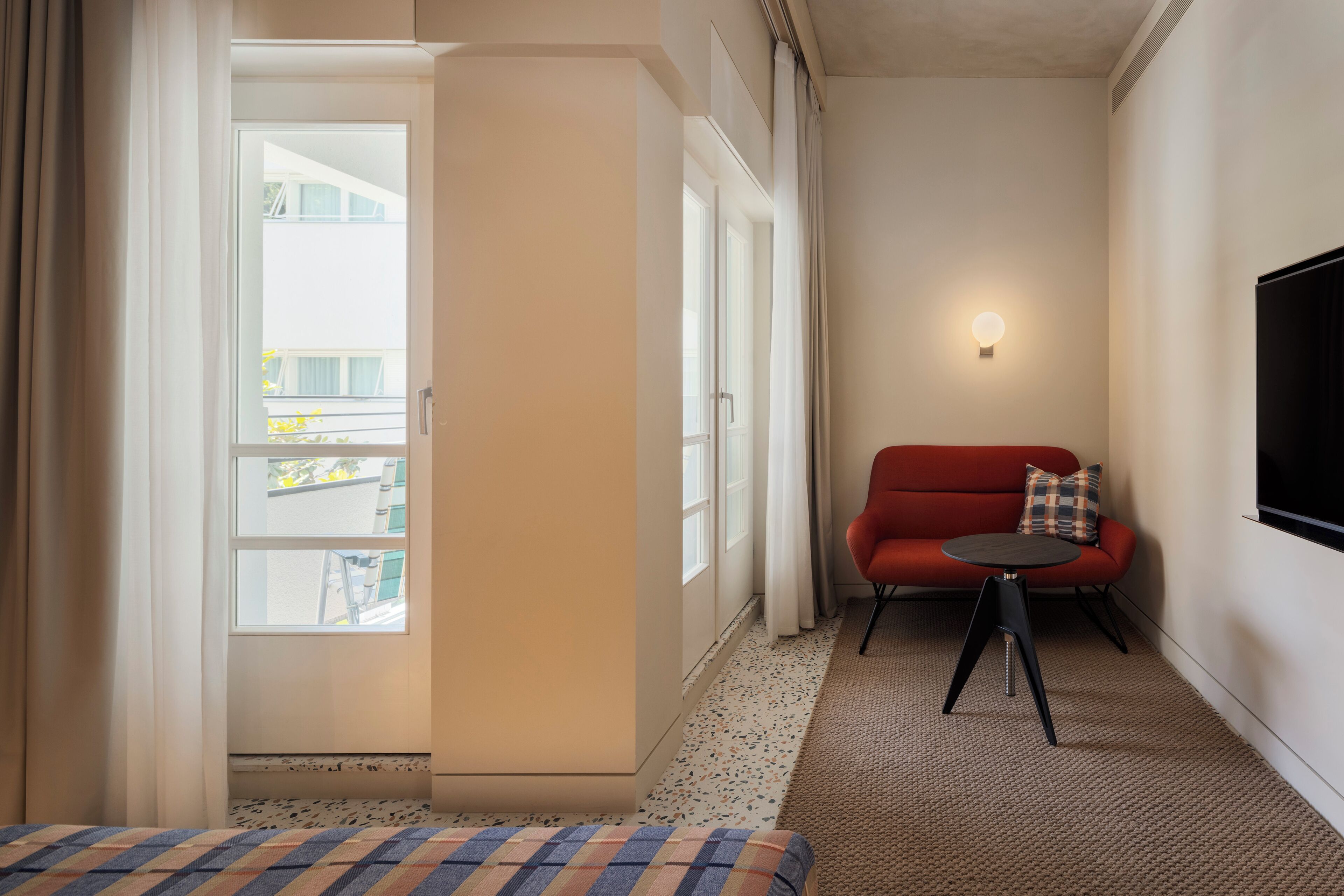 Une chambre d’hôtel avec un lit, un canapé rouge, une petite table, un téléviseur fixé au mur et une fenêtre avec des rideaux.