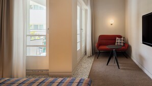 Une chambre d’hôtel avec un lit, un canapé rouge, une petite table, un téléviseur fixé au mur et une fenêtre avec des rideaux.