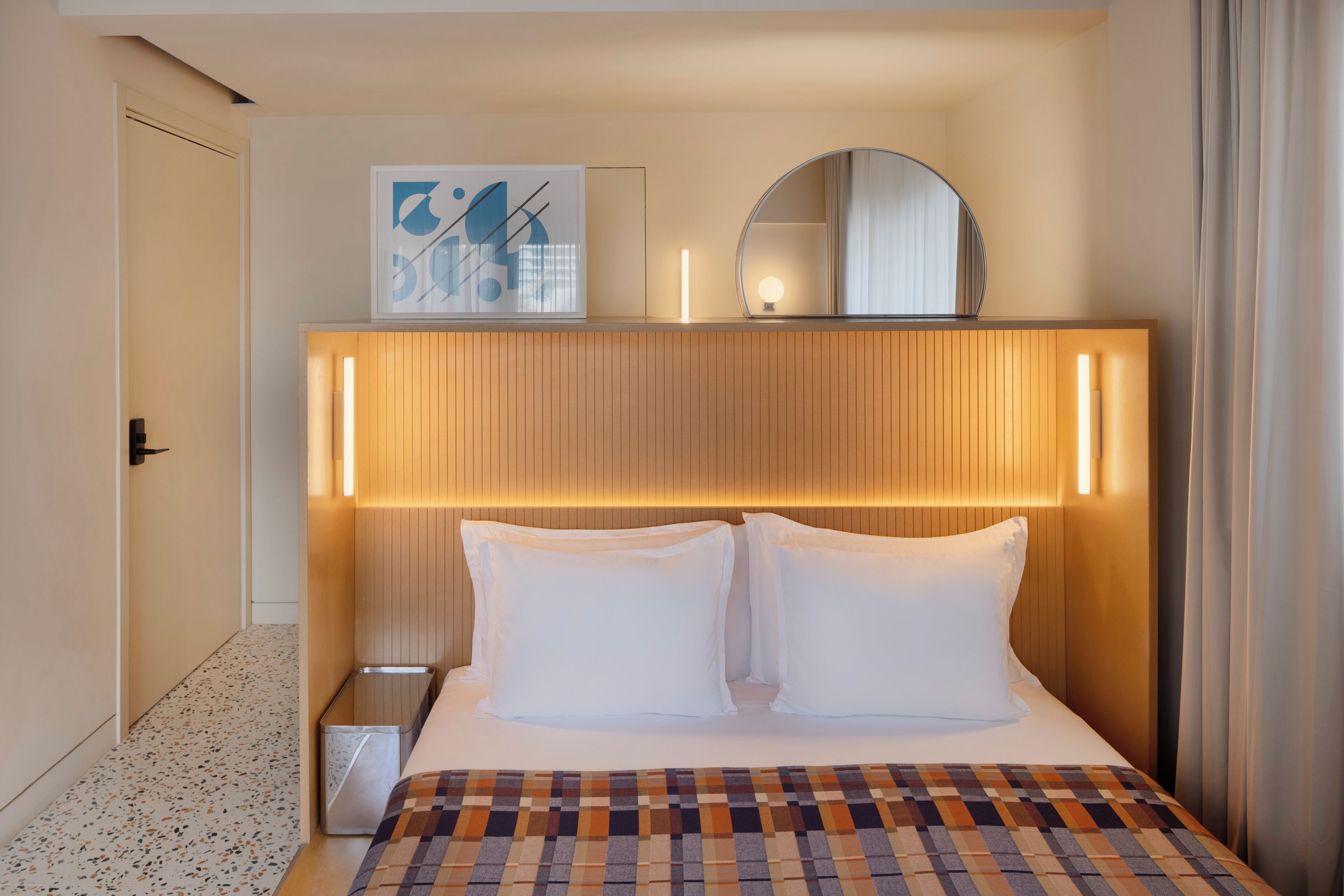 A hotel room with a bed, a bedside table, a wall-mounted mirror, and a framed picture.