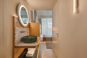 Un bagno con un lavandino tondo, uno specchio illuminato, un portascopino e vista su un balcone.
