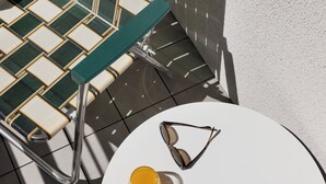 Une chaise avec un siège et un dossier en osier tressé, une paire de lunettes de soleil et un verre de jus d’orange sur une table blanche.