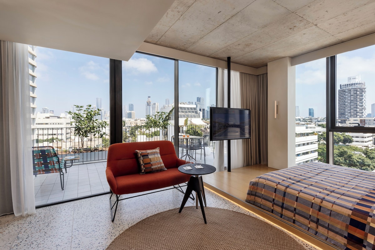 Camera d'albergo moderna con un letto grande, un divano rosso, una TV e un balcone con vista sulla città.