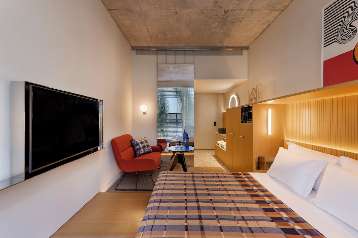 Una camera d'albergo moderna con un letto grande, una TV a schermo piatto, una poltrona rossa, un piccolo tavolo da pranzo e un armadio.