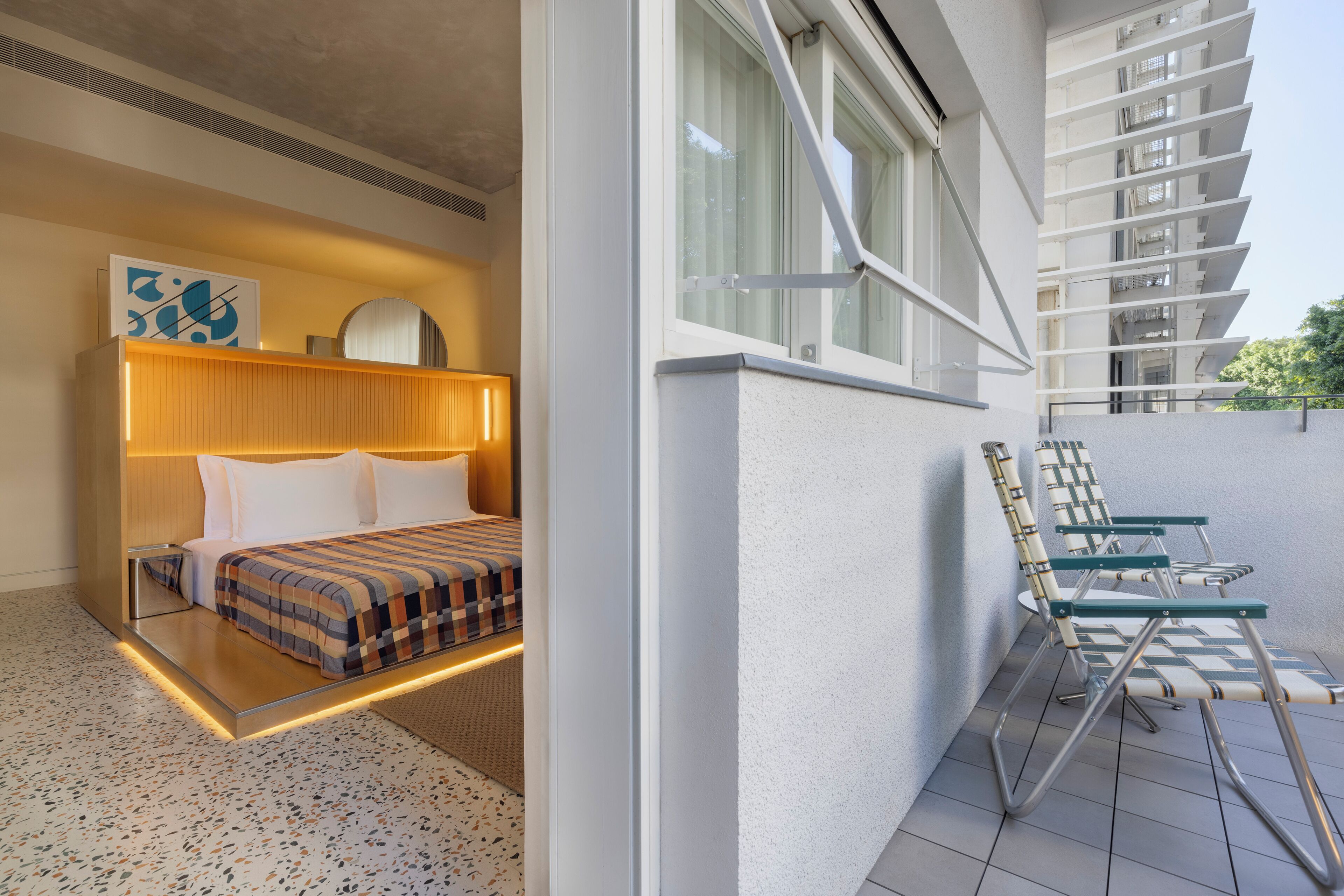 Une chambre d’hôtel moderne avec un lit, une table de chevet et une vue sur un balcon équipé de chaises.