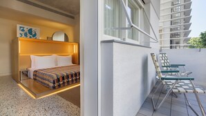 A modern hotel room with a bed, bedside table, and a view of a balcony with chairs.