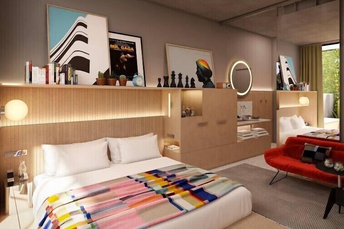 Una camera d'albergo moderna con un letto grande, un divano rosso, una testiera in legno e una libreria con libri e oggetti decorativi.