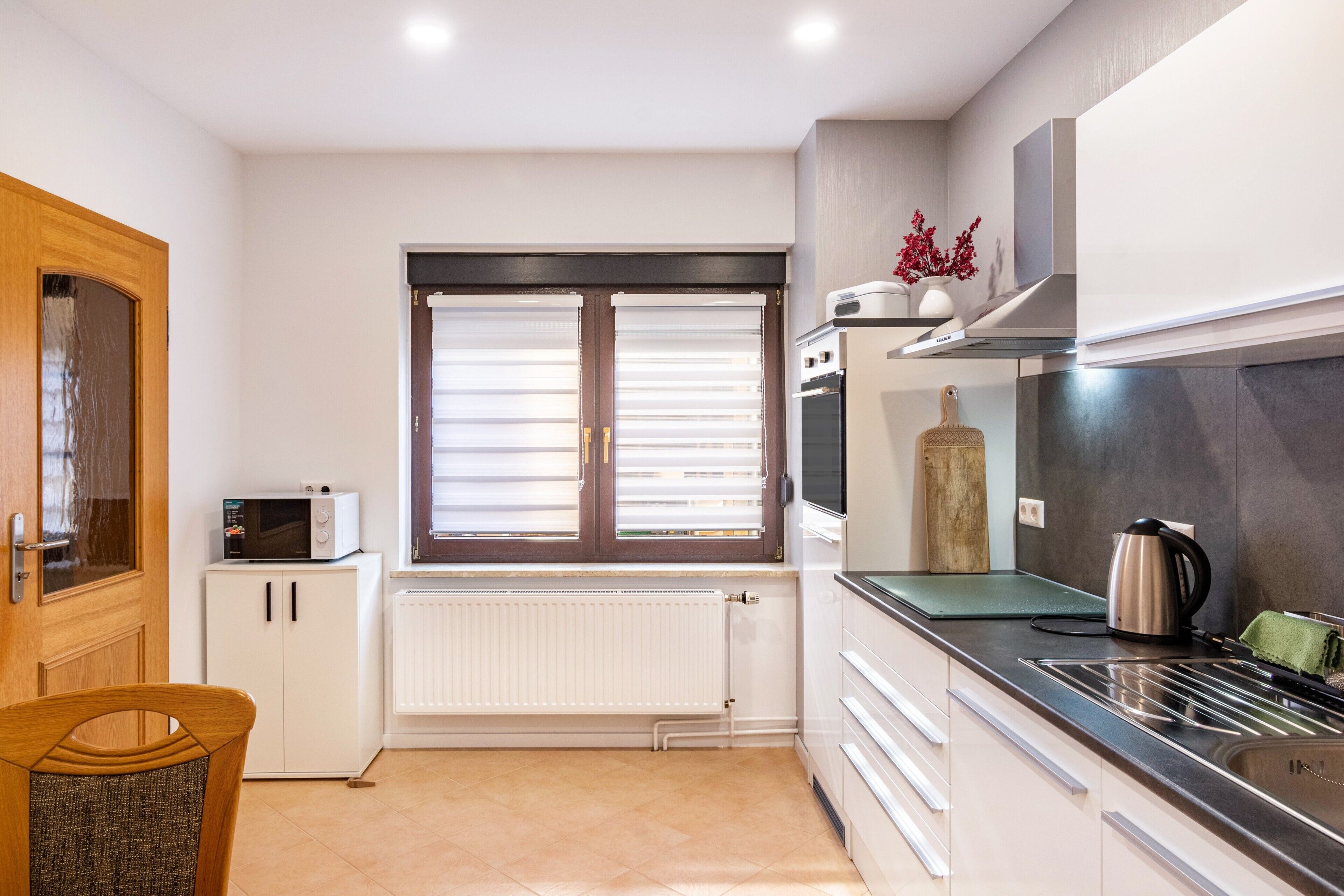 A modern kitchen with a built-in microwave, oven, and stove. There is a window with blinds, a radiator, and a wooden door.