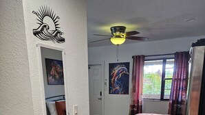 Quarto com cama, ventilador de teto, janela com cortinas, porta e parede com mural de sol e ondas.