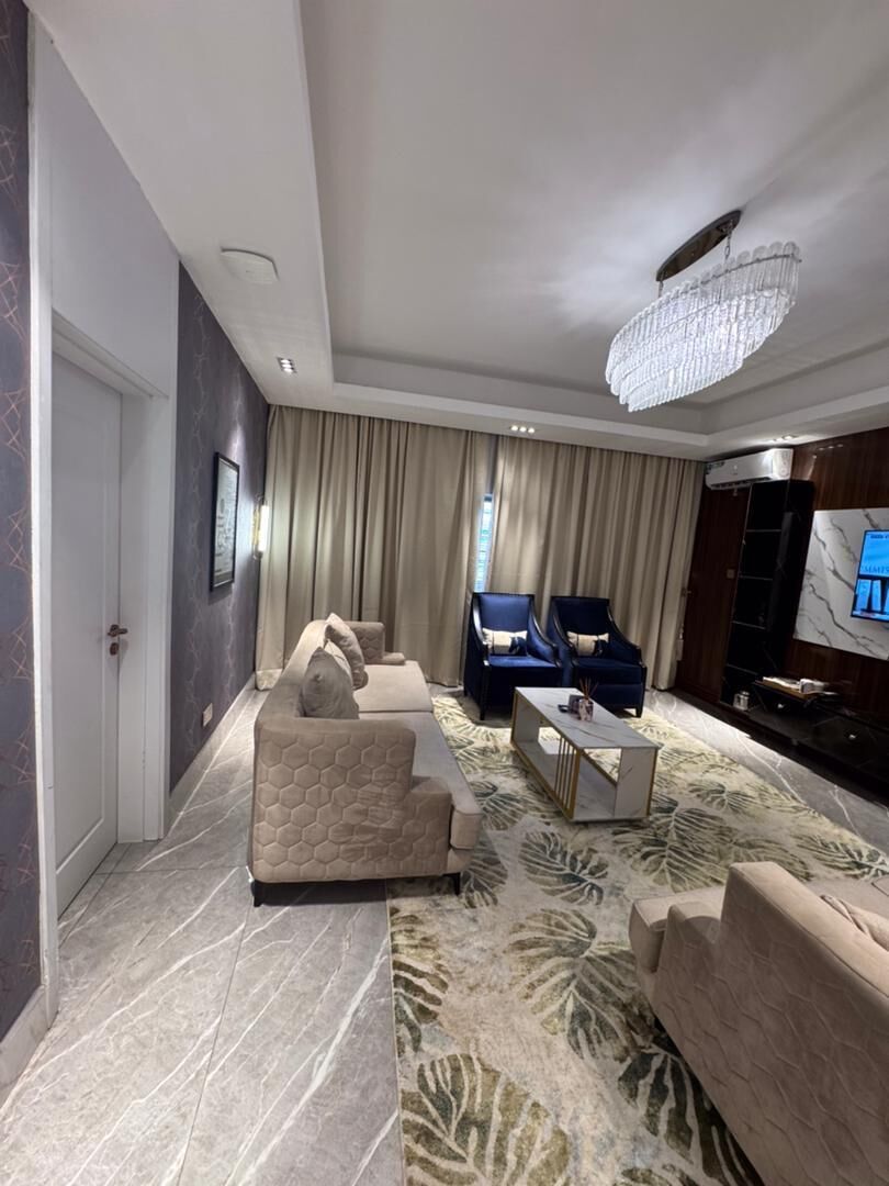A modern living room with a sofa set, two armchairs, a coffee table, a chandelier, and a flat-screen TV.