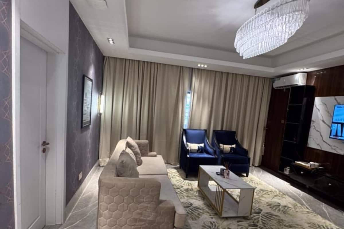 A modern living room with a sofa set, two armchairs, a coffee table, a chandelier, and a flat-screen TV.