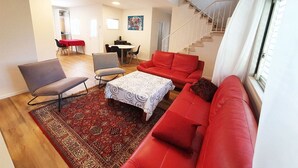 Ein geräumiger Wohnbereich mit einem roten Sofa, einem gemusterten Hocker und einem Esstisch mit Stühlen. Im Hintergrund ist eine Treppe zu sehen.